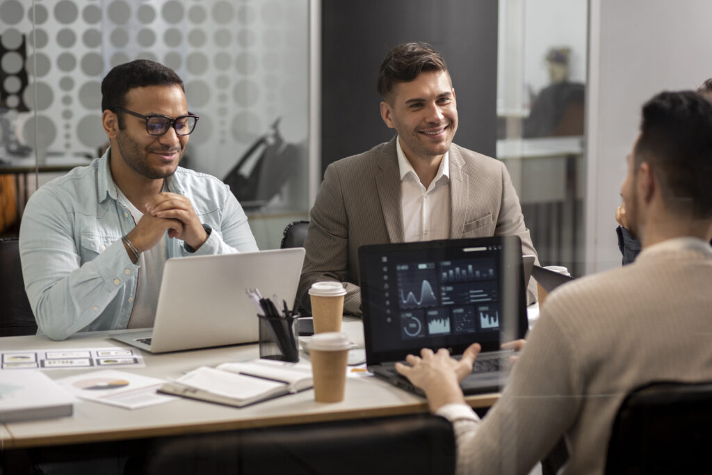 Três homens em uma reunião de negócios, discutindo dados exibidos em um laptop, em um ambiente corporativo moderno.