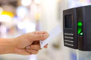 Pessoa aproximando um cartão de acesso branco a um leitor eletrônico com teclado numérico e luz verde acesa.
