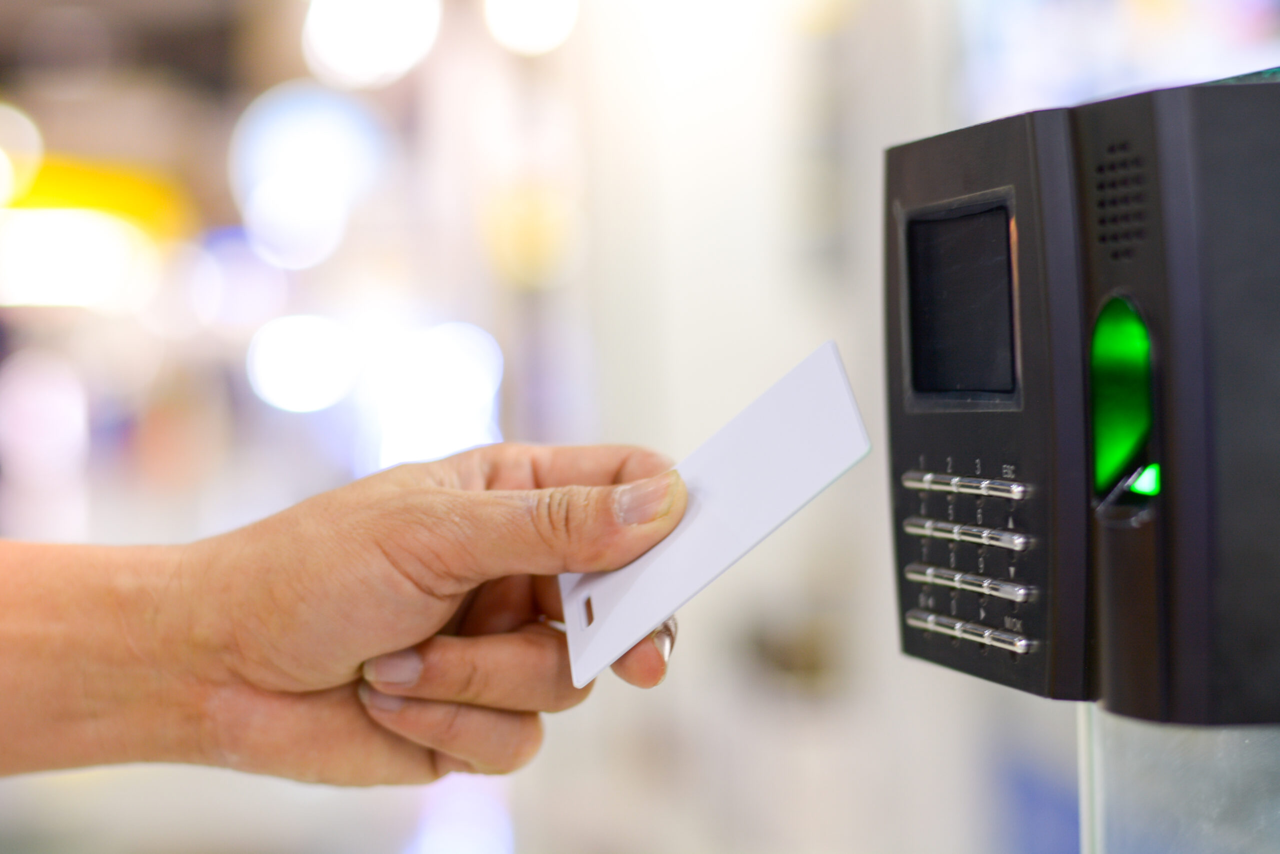 Pessoa aproximando um cartão de acesso branco a um leitor eletrônico com teclado numérico e luz verde acesa.