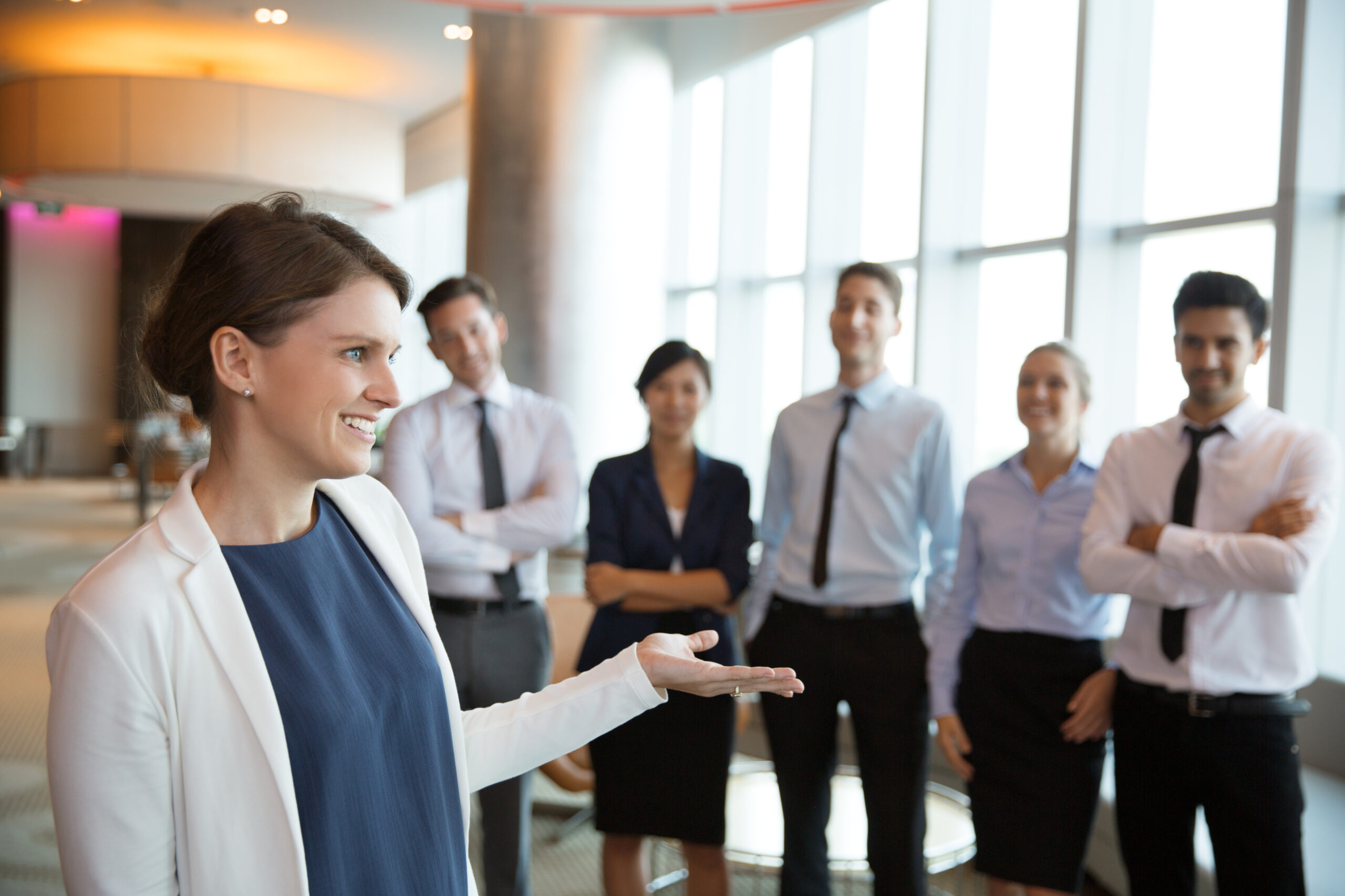 ulher sorridente em destaque, apresentando ou interagindo com uma equipe de profissionais em um ambiente corporativo moderno.