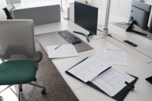 Mesa de escritório com computador desligado, documentos espalhados e cadeira vazia.