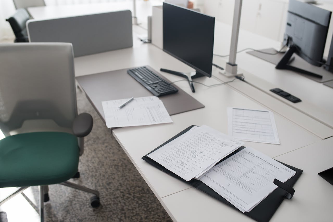 Mesa de escritório com computador desligado, documentos espalhados e cadeira vazia.