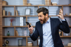 Homem de terno irritado grita com o celular na mão, demonstrando frustração em ambiente de escritório.
