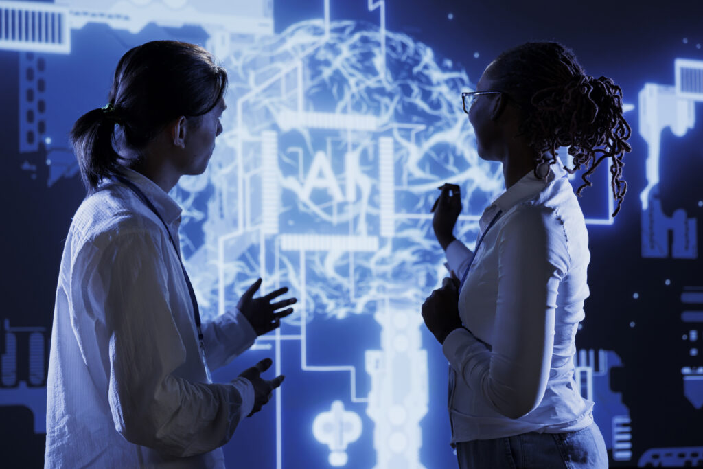 Duas pessoas de perfil corporativo analisam uma projeção digital com o desenho de um cérebro e o termo “IA” ao centro, simbolizando inteligência artificial.
