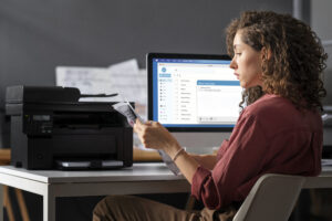 Mulher sentada em escritório analisa documentos impressos ao lado de uma impressora, com e-mail aberto em monitor ao fundo.