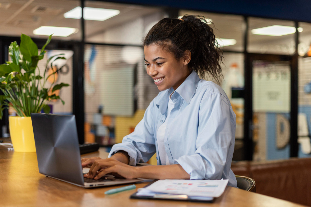 Mulher sorridente usando camisa azul clara digitando em um notebook, sentada em uma mesa com documentos e uma caneta, em um espaço de coworking.