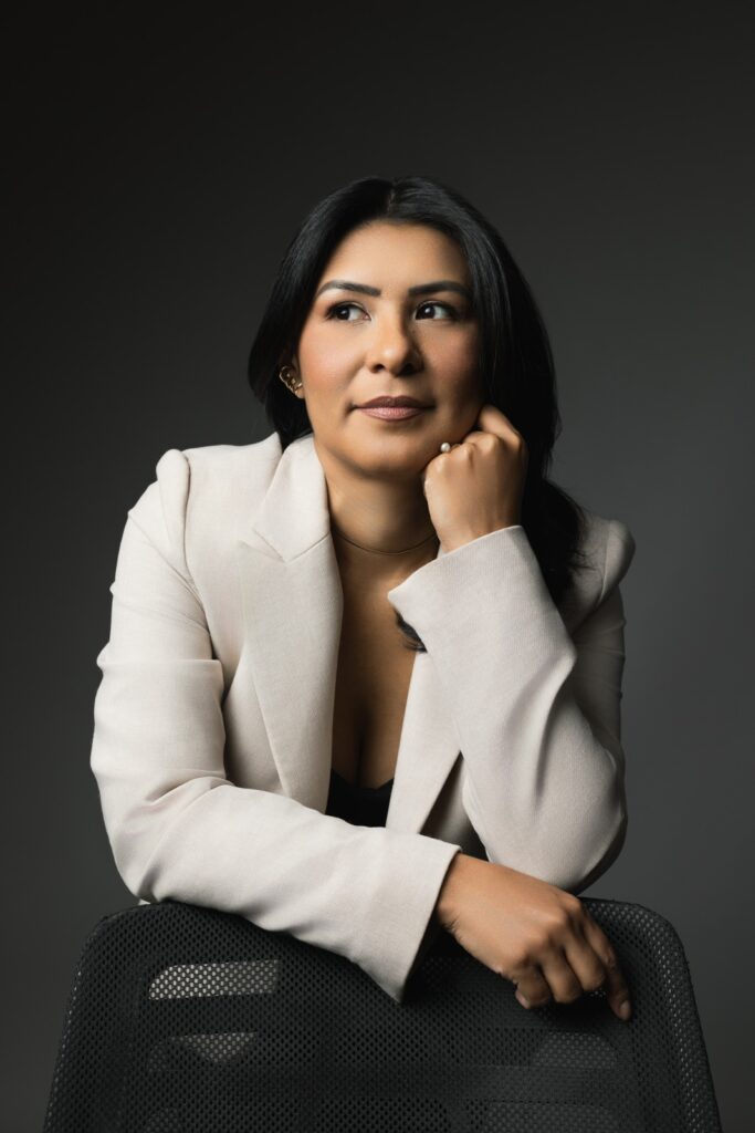 Mulher com blazer claro apoiada em encosto de cadeira preta, olhando pensativa para o lado.