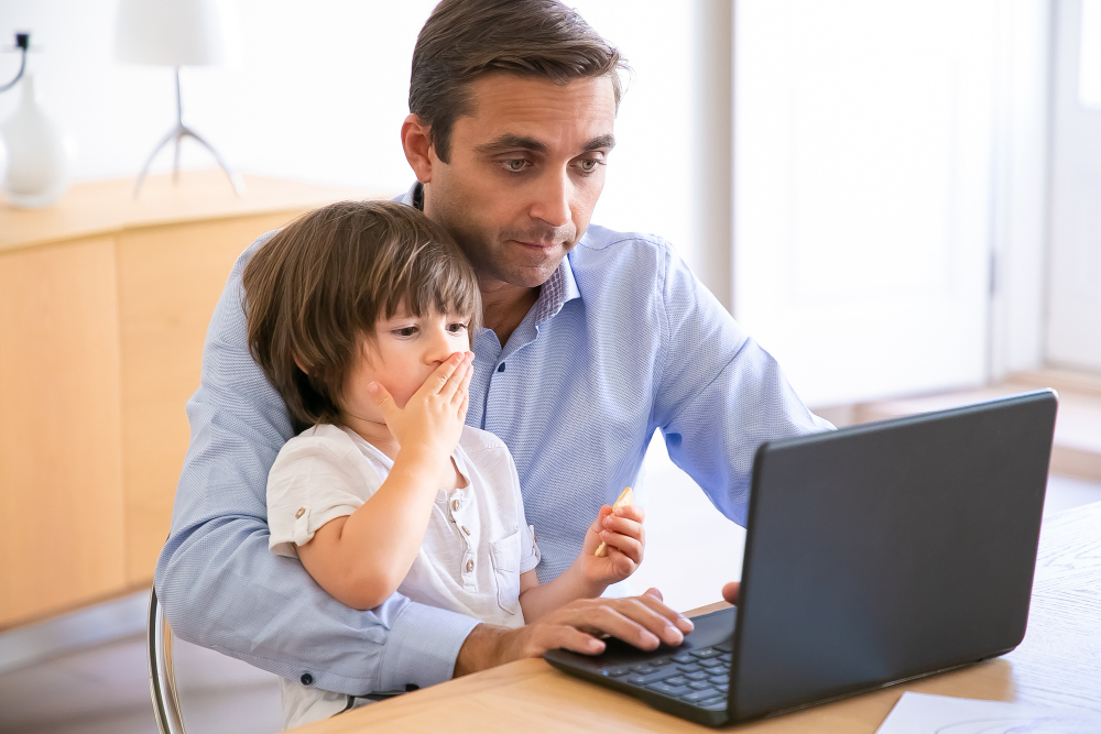 Homem sentado à mesa, usando um notebook, com uma criança pequena no colo. O pai olha para a tela enquanto a criança leva a mão à boca, ambos concentrados.