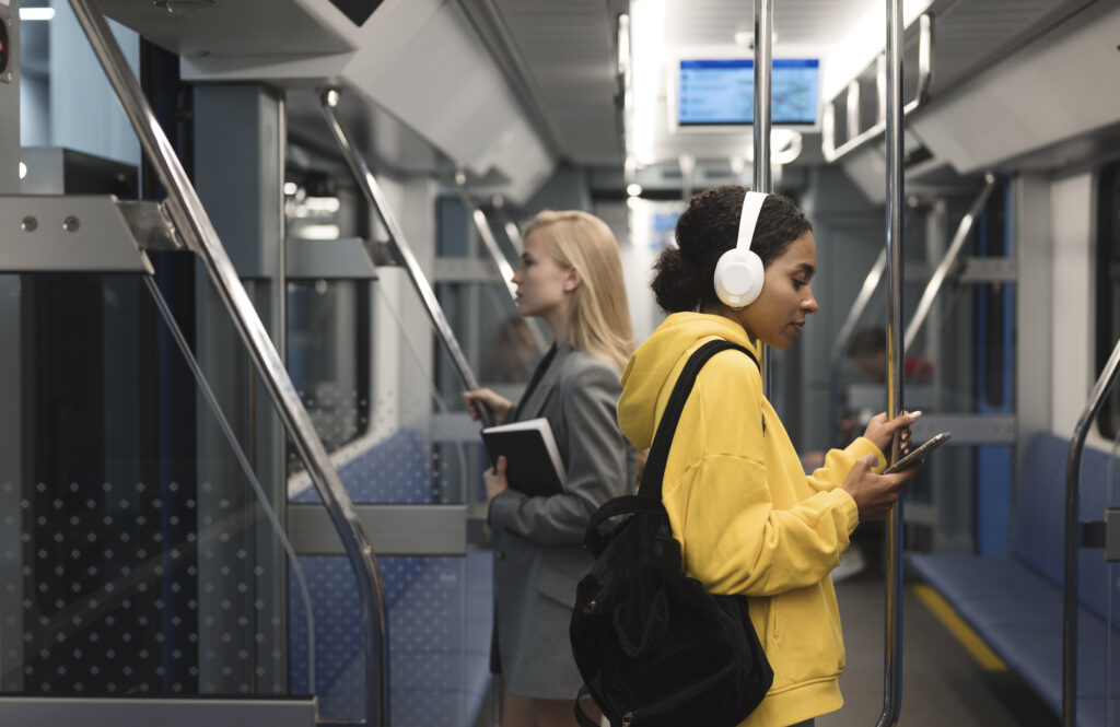 Duas pessoas viajam de metrô; uma jovem de moletom amarelo com fones de ouvido usa o celular, enquanto outra segura um livro e observa pela janela
