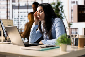Mulher em escritório com expressão de estresse, mão na testa, em frente a laptop. Ilustra o esgotamento e a insatisfação no trabalho.