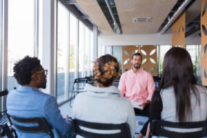 Quatro pessoas sentadas em cadeiras em uma sala iluminada, participando de uma conversa em grupo, com um facilitador à frente conduzindo a discussão.
