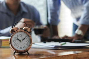 Relógio despertador em uma mesa de escritório com duas pessoas analisando documentos ao fundo, representando o controle de horas extras no ambiente de trabalho.