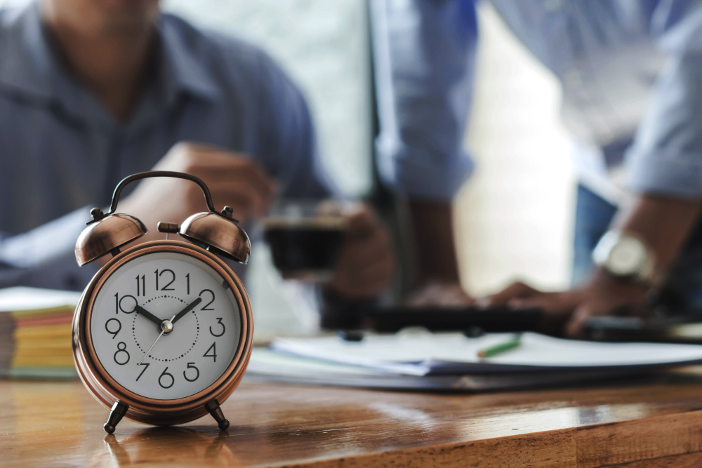 Relógio despertador em uma mesa de escritório com duas pessoas analisando documentos ao fundo, representando o controle de horas extras no ambiente de trabalho.