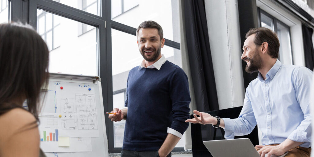 Grupo de profissionais em uma reunião de negócios. Um homem em pé sorrindo, apresenta um gráfico em um quadro branco para três colegas.