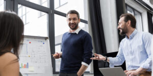 Grupo de profissionais em uma reunião de negócios. Um homem em pé sorrindo, apresenta um gráfico em um quadro branco para três colegas.