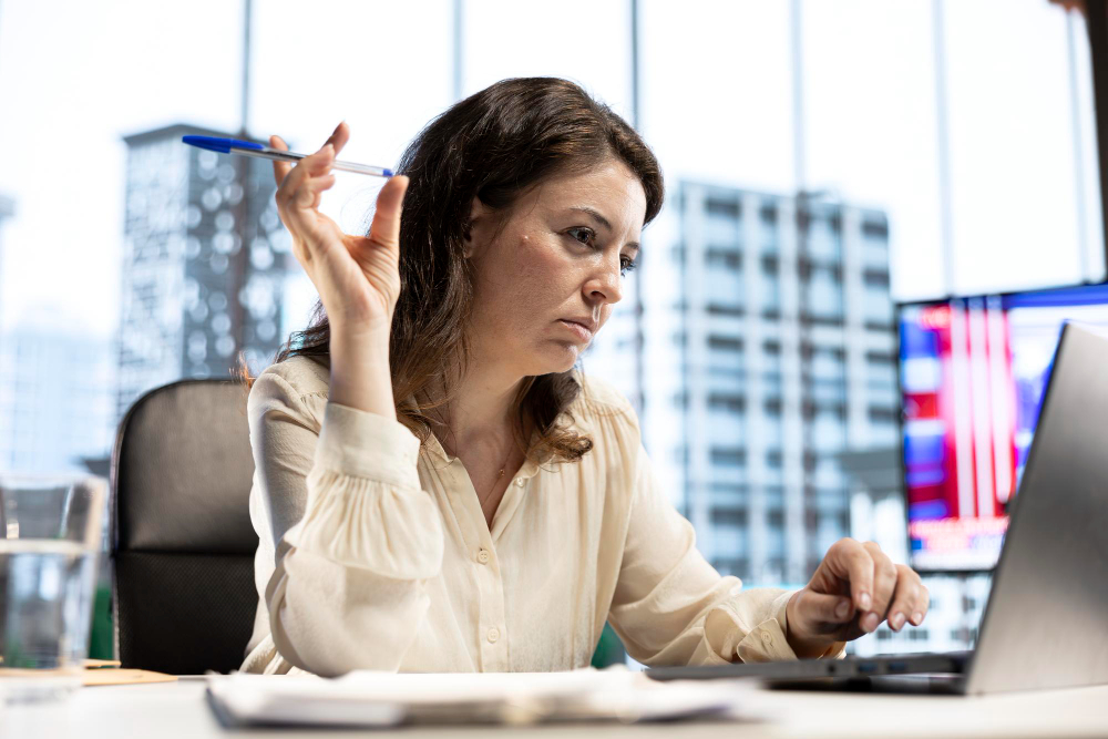 Profissional concentrada trabalhando no notebook em escritório, segurando uma caneta durante análise de dados.
