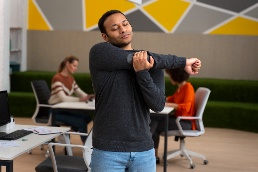 Colaborador alongando os braços em ambiente de escritório enquanto outros profissionais trabalham ao fundo.