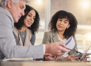 Três profissionais analisando documentos e conversando em reunião de trabalho, com laptop e papéis sobre a mesa.