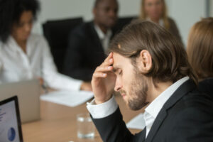 Homem em um ambiente corporativo, sentado durante uma reunião, com expressão de cansaço ou preocupação.