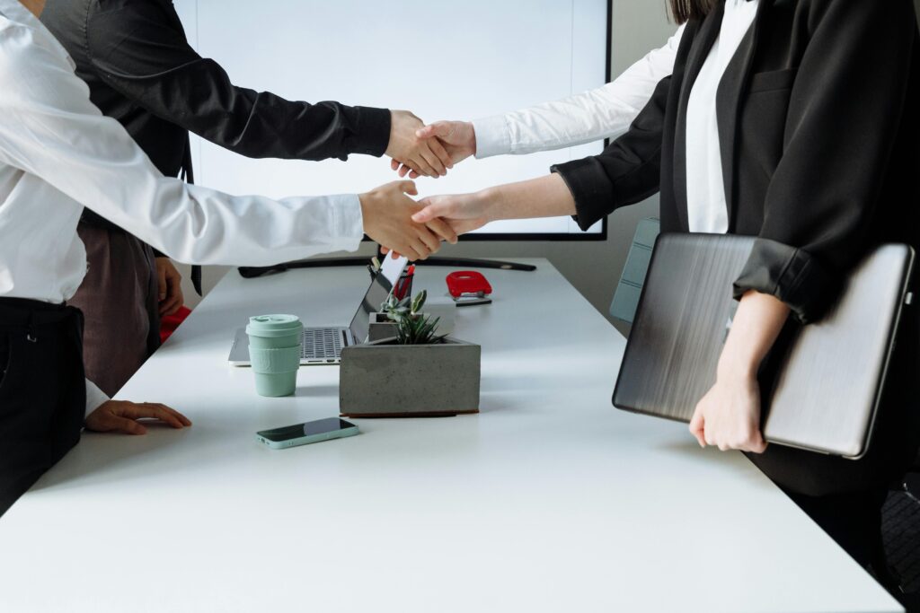 Duas pessoas apertando as mãos em uma reunião de negócios, com outras pessoas ao redor de uma mesa de escritório.