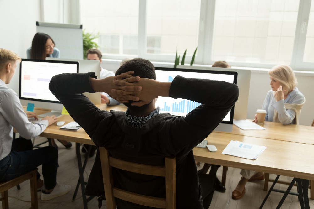 Equipe reunida em escritório durante reunião de trabalho, com homem sentado de costas e mãos atrás da cabeça observando gráficos em monitores.