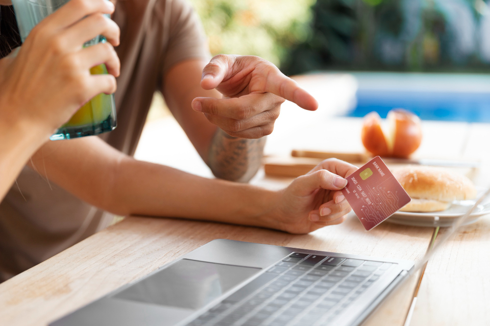 Foto em plano detalhado e ângulo diagonal de duas pessoas sentadas a uma mesa de madeira ao ar livre. Uma delas segura um cartão de benefícios na cor vermelha sobre um notebook aberto, enquanto a outra, ao fundo, aponta o dedo em direção à tela. Em primeiro plano, do lado esquerdo, aparecem as mãos de uma pessoa segurando um copo com suco amarelo. Sobre a mesa, há também um prato com um sanduíche e, ao fundo, a área azul de uma piscina desfocada.