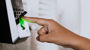 Foto em close-up de um colaborador encostando o dedo indicador no leitor de um relógio de ponto biométrico. O leitor brilha com uma luz verde ao validar a biometria, mantendo o fundo do escritório desfocado.