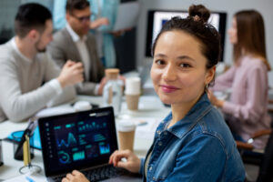 Uma profissional sorridente em um escritório moderno, sentada com um laptop aberto que exibe dashboards e gráficos de análise de dados (representando o uso de ferramentas de IA), com colegas em reunião desfocados ao fundo.