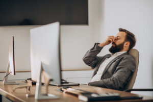 Homem trabalhando em frente ao computador, com expressão de cansaço e estresse, representando os impactos do retrabalho no ambiente corporativo.