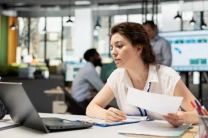 Profissional analisa documentos em uma mesa de escritório, com um notebook e outros materiais de trabalho ao redor.
